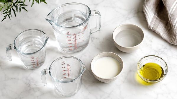 Glass measuring jugs and bowls of water, milk, and olive oil on white marble — cups to ml converter tool
